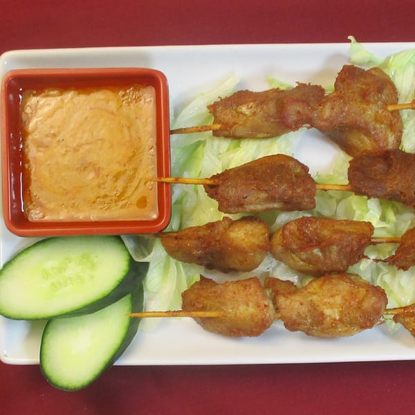Thai chicken satay skewers with peanut sauce and cucumber slices on a white plate.